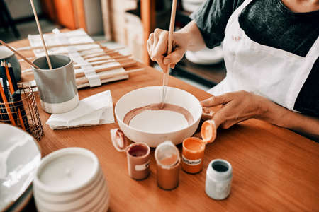 I use art to escape. Cropped shot of an unrecognizable woman sitting alone and painting a pottery bowl in her workshop.の写真素材
