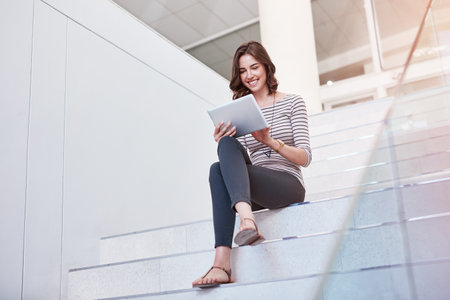 Prompt responses make for great email etiquette. Shot of a young businesswoman using a digital tablet on the stairs in a modern office.の写真素材