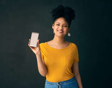 Check out this amazing mobile app. Shot of a young woman holding a cellphone while posing against a grey background.の写真素材