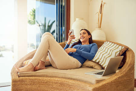 Want to meet up for coffee later. Shot of a happy young woman using her smartphone while relaxing in...