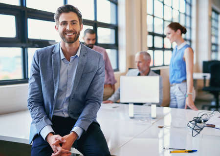 Hes the face of job satisfaction. Portrait of a confident businessman posing in the office with his colleagues in the background.の写真素材