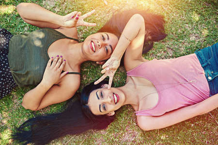 Life is all good. Cropped shot of two young friends showing the piece sign while lying on the grass.