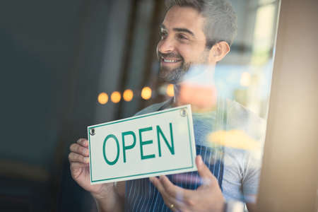 Time to serve some awesome coffee. Shot of a handsome young man hanging up an open sign on the door...