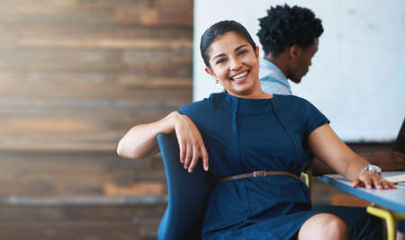 Having a great day at work. A young businesswoman sitting in an office chair with a colleague in the background.の写真素材