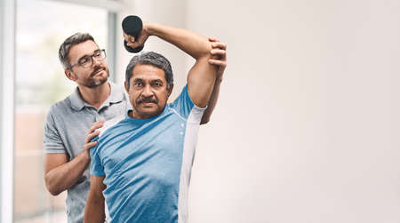 Proper technique is the main focus. Shot of a senior man exercising with dumbbells during a rehabilitation session with his physiotherapist.の写真素材
