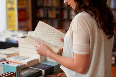 Lost in the pages. A cropped shot of a young woman reading a book while standing in a bookstore.の写真素材