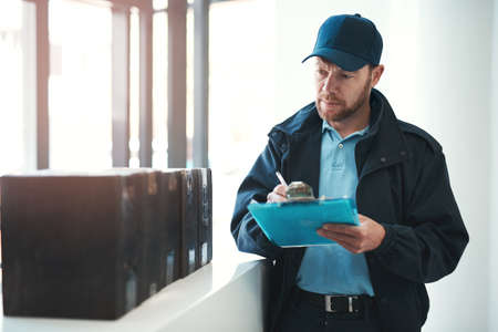 Making sure every delivery is signed and dated. Shot of a handsome delivery man writing on his clipboard while waiting in the lobby with a customers order.の写真素材