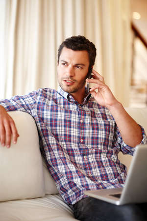 Working from home. A handsome man speaking on his cellphone while sitting with his laptop on the...