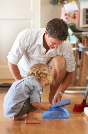 They make a terrific team. A father holding a dustpan while his toddler son sweeps sand into it using a hand broom..の写真素材
