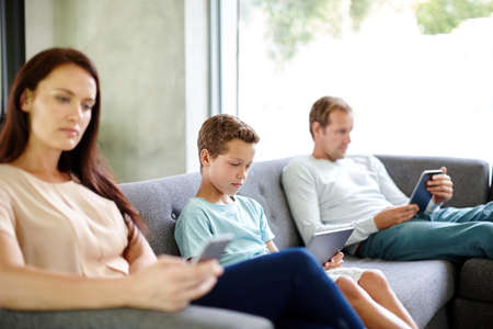 Left to their own devices.... Shot of a family of three sitting separately on a sofa with their own digital devices.の写真素材