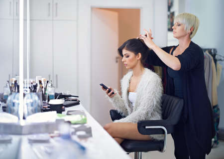 Looking gorgeous sometimes requires a bit of help. A young model at the hair salon having her done while checking her phone.の写真素材