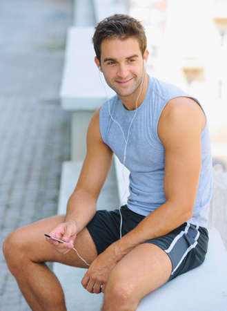 Exercise invigorates him. Happy young jogger sitting outside and listening to music.