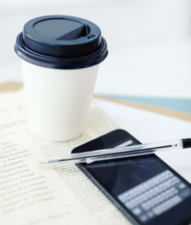 Coffee and the crossword. Image of a smartphone on a newspaper with a pen on top of it, and a disposable coffee cup.の写真素材