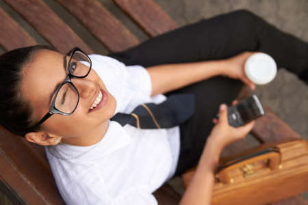 I learn, what is your superpower. A beautiful young woman winking at the camera while sitting on a bench with her cellphone, coffee and satchel.の写真素材