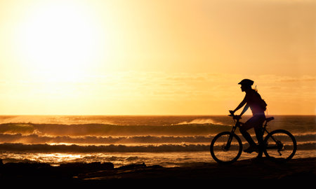 Cycling off into the sunset. A silhouette of a female cyclist on the beach at sunset.の写真素材