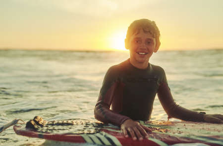 You cant be sad when youre on a surfboard. Portrait of a young boy in a wetsuit sitting on his surfboard in the ocean.の写真素材