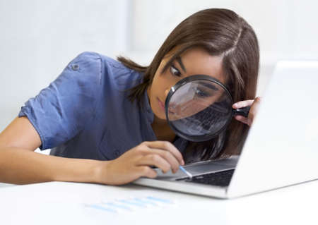 Not missing a speck of dirt.... Young woman using a magnifying glass while cleaning her laptop...