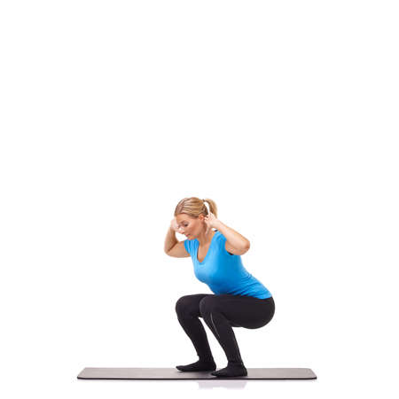 Doing squats with arms raised. A young woman crouching on her exercise mat while isolated on a white background.の写真素材