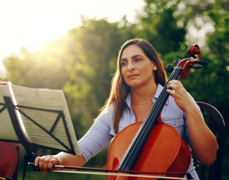 Music is something you hear and feel. Cropped shot of a beautiful woman playing a cello in the backyard.の写真素材