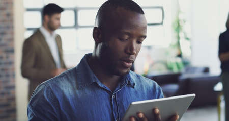 Trying out his new personal helper. Cropped shot of an handsome young businessman working on his digital tablet with a few colleagues in the background.の写真素材