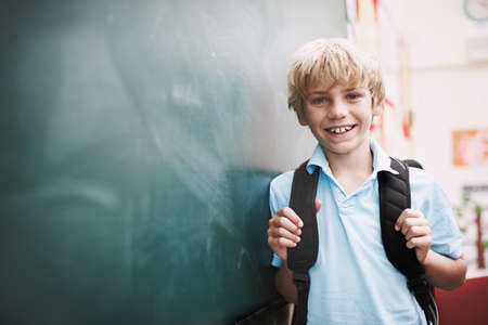 Hes ready to learn. An happy young boy standing alongside copyspace at the blackboard and holding the straps of his backpack.の写真素材