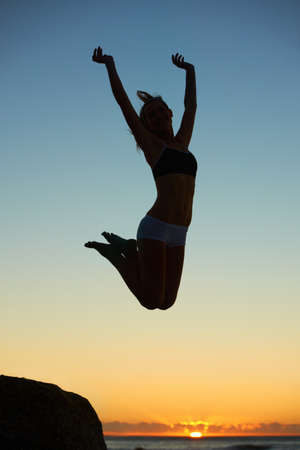Jumping for fun. Silhouette of a young woman jumping in the air at sunset.の写真素材