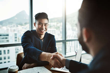 Its safe to say he made a great impression. Shot of two young businessmen shaking hands while sitting at a desk in a modern office.の写真素材