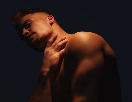 Im a handsome man with a charming personality. Shot of a handsome young man with no shirt on standing in the shadows.の写真素材