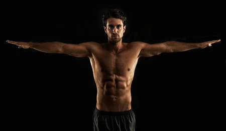 Perfect form. Handsome topless man raising his arms to his side - Isolated on black.