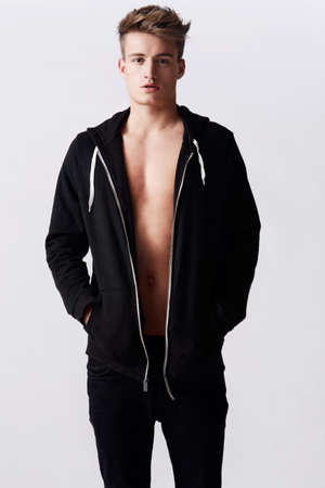 Male fashion model. Studio portrait of young attractive man wearing a hoodie standing with hands in...