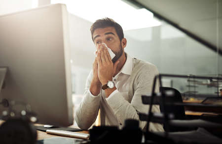 Could this be the flu coming on. Shot of a handsome young businessman blowing his nose while working late in an office.の写真素材