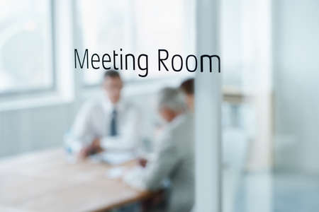 The meeting is in session. Closeup of a glass door reading meeting room with business people blurred...
