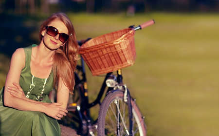 Perfect day for a bike ride in the countryside. Shot of a beautiful young woman relaxing in the park next to her old fashioned bicycle.の写真素材