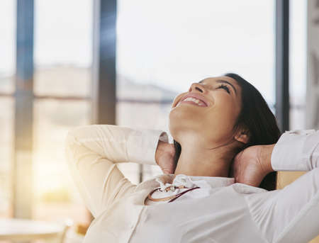 Owning her success. Shot of a content young businesswoman sitting in her office with her hands behind her head.の写真素材
