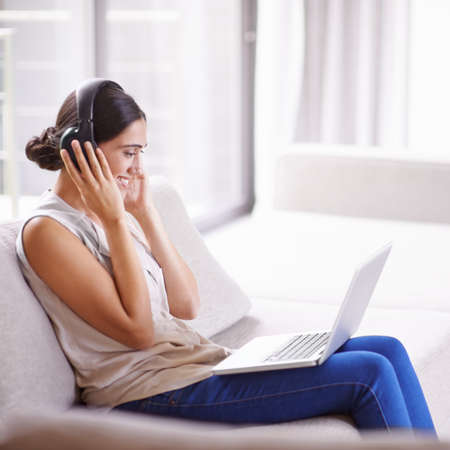 Staying in touch in the digital age. Shot of a young woman on a sofa wearing headphones and using a...