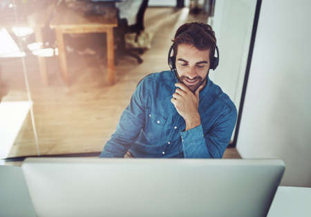 Hes always happy to be of assistance. High angle shot of a handsome young businessman wearing a headset while sitting at his desk.の写真素材