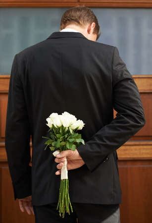 Hes a man of mystery. A man wearing a suit holding a bunch of white roses behind his back.の写真素材