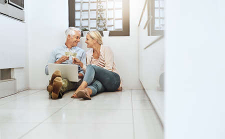 Heres to us, babe. Full length shot of an affectionate mature couple drinking wine while browsing the web on their kitchen floor.の写真素材