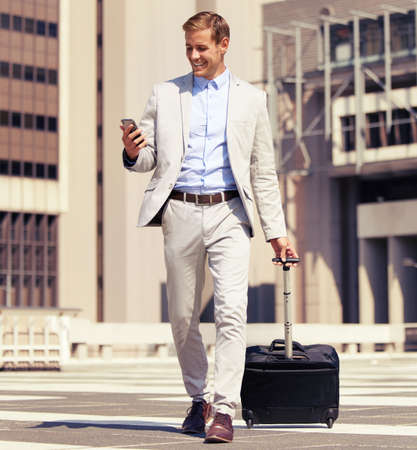 This app is convenient for a businessman on the go. Shot of a young businessman using a cellphone while walking with a suitcase in the city.の写真素材