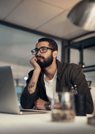 Better finish up before I fall asleep. Shot of a young businessman looking uncertain while working late at night in a modern office.の写真素材