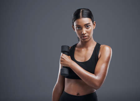Watch me get stronger. Studio portrait of a sporty young woman lifting weights against a grey background.の写真素材