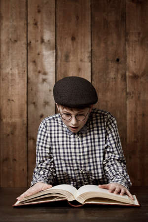 I cant believe what Im reading. Young boy in retro clothing wearing spectacles reading a massive book with a surprised expression.の写真素材