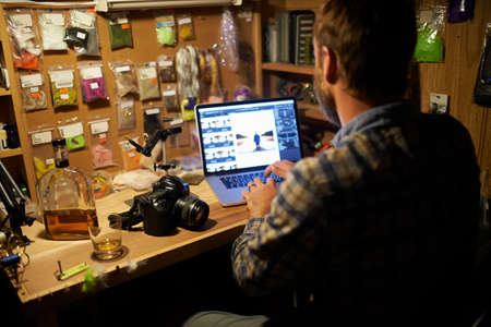 Downloading some photographs. Shot of a man using his laptop while working in his fly fishing workshop.の写真素材