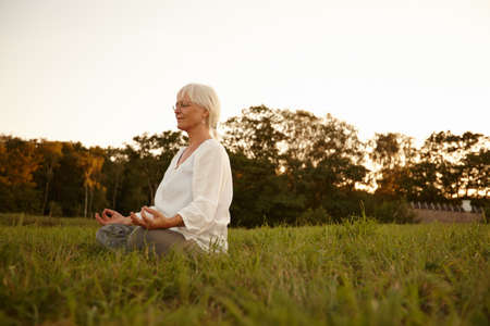 Meditation in the meadows. Shot of an attractive mature woman meditating in the outdoors at sunset.の写真素材