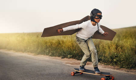Ready, steady, fly. Shot of a young boy pretending to fly with a pair of cardboard wings while riding a skateboard outside.の写真素材