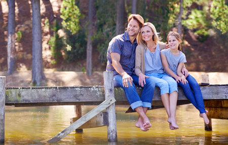 Family time is so precious. A happy family of three sitting together on the jetty at the lake.の写真素材