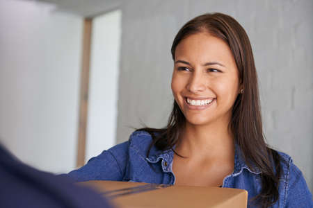 Excited to get settled in. A beautiful young woman smiling at the camera as she takes a box from a mover.の写真素材