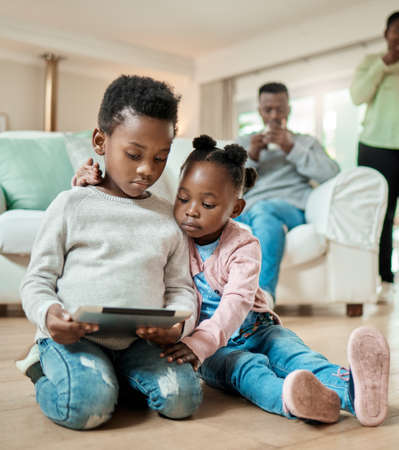 Shes always interested on what her brothers doing. Full length shot of young boy and his adorable little sister using a tablet while sitting on the living room floor at home.の写真素材