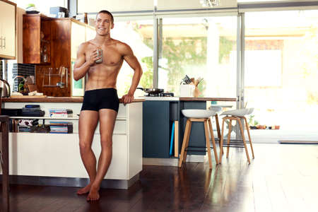 I love my time off just to enjoy a coffee. Shot of a handsome young shirtless man drinking a cup of coffee in the kitchen at home.の写真素材