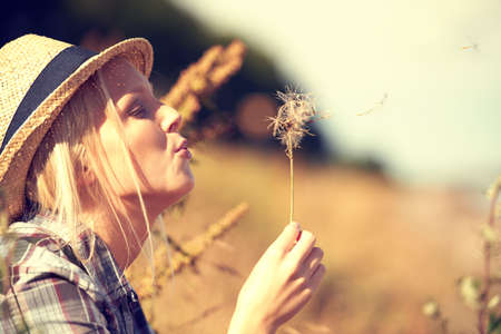 Blowing away her cares.... Beautiful young woman wearing a straw fedora and blowing at a dandelion.の写真素材
